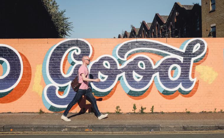 an orange wall on the side of a street. a person walks left to right in front of the wall on a sidewalk. the wall has dark blue graffitied typography that says "good"