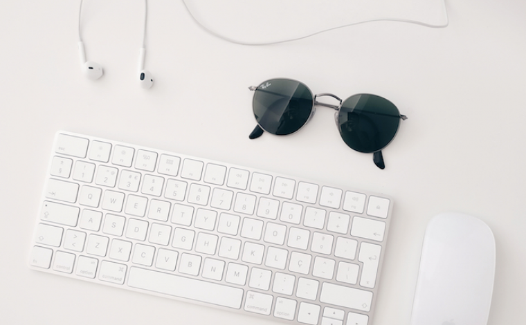 overhead image of a white keyboard, white mouse, white wired headphones, and a pair of black sunglasses