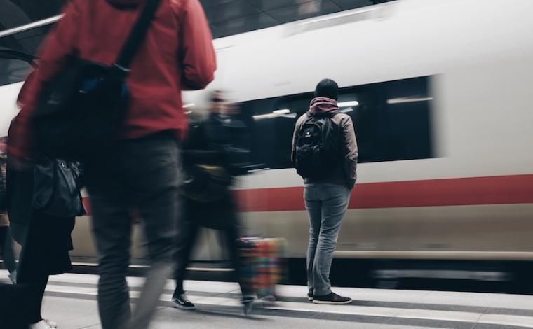 a person wearing a black backpack waits for a train as it whizzes by them. in the unfocused shot of the photo, 3 others wait for the train or walk past